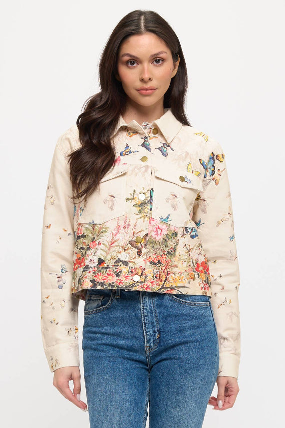 A woman with long brown hair models the Butterfly Floral Jacket, showcasing colorful florals and butterflies, paired with blue jeans against a plain white background.