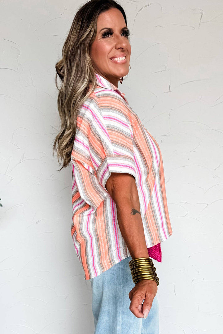 A woman with long wavy hair smiles while standing sideways. She wears the Multicolor Stripe Shirt in pink, orange, and white, styled with light blue jeans, gold bangles, and displays a small tattoo on her forearm.