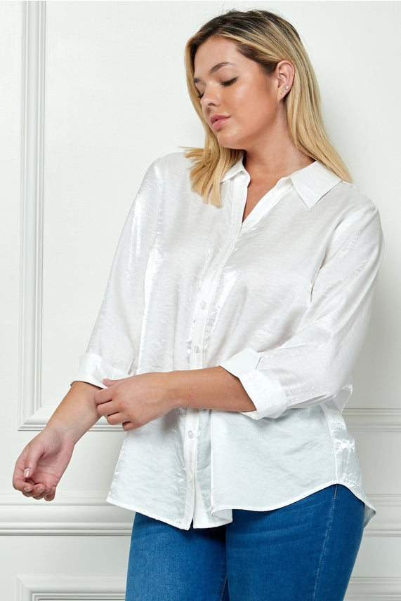 A woman with blonde hair models the Plus Shimmer Collar Blouse, a white shimmery top with rolled-up sleeves, paired with blue jeans. She stands against a white wall, gazing down and appearing relaxed.