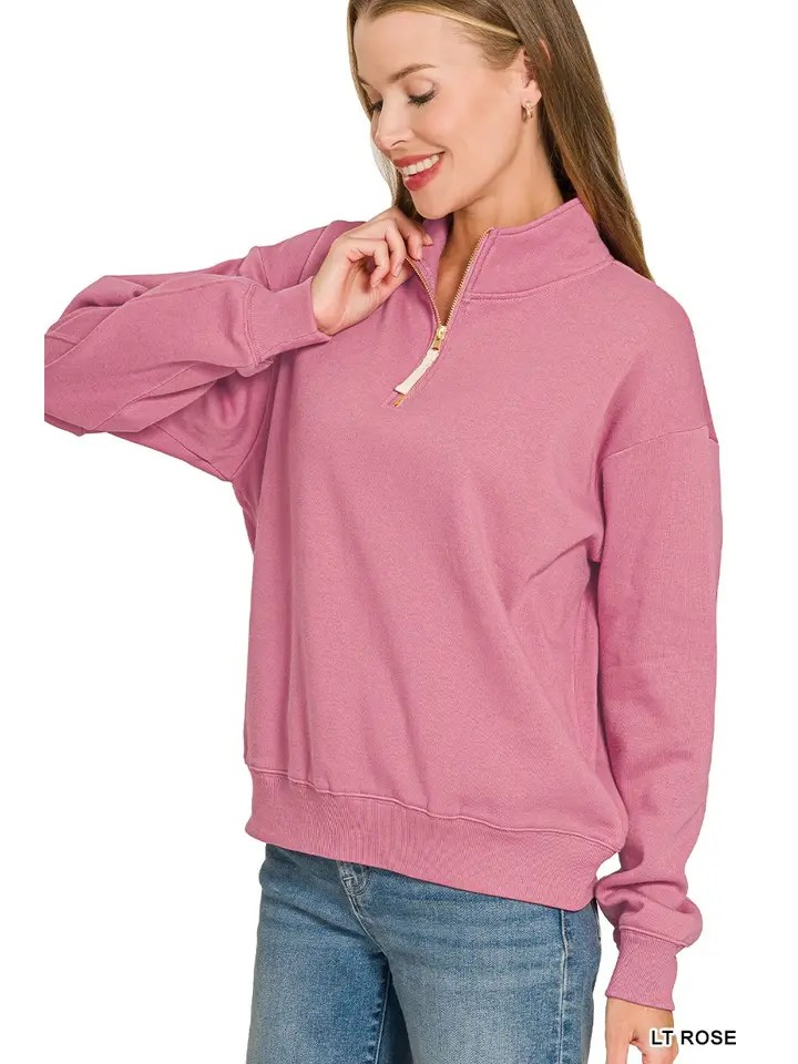 A woman stands and smiles with one hand touching her chin, wearing the Fleece Half Zip Sweatshirt in light rose with a relaxed fit, paired with blue jeans.