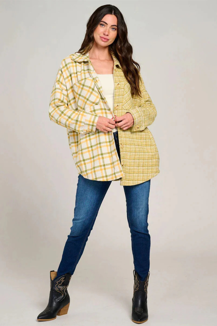 A full-body shot of a woman with long brown hair, wearing an unbuttoned yellow, white, and green plaid shacket over a white top, blue skinny jeans, and black cowboy boots, standing with one leg slightly bent.