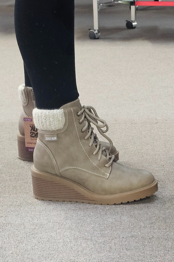 A person wearing tan Sky Solange Wedge Boots with knit cuffs stands on a beige carpet. The faux leather boots have thick rubber soles and are styled with black leggings. A wheeled chair appears in the background.