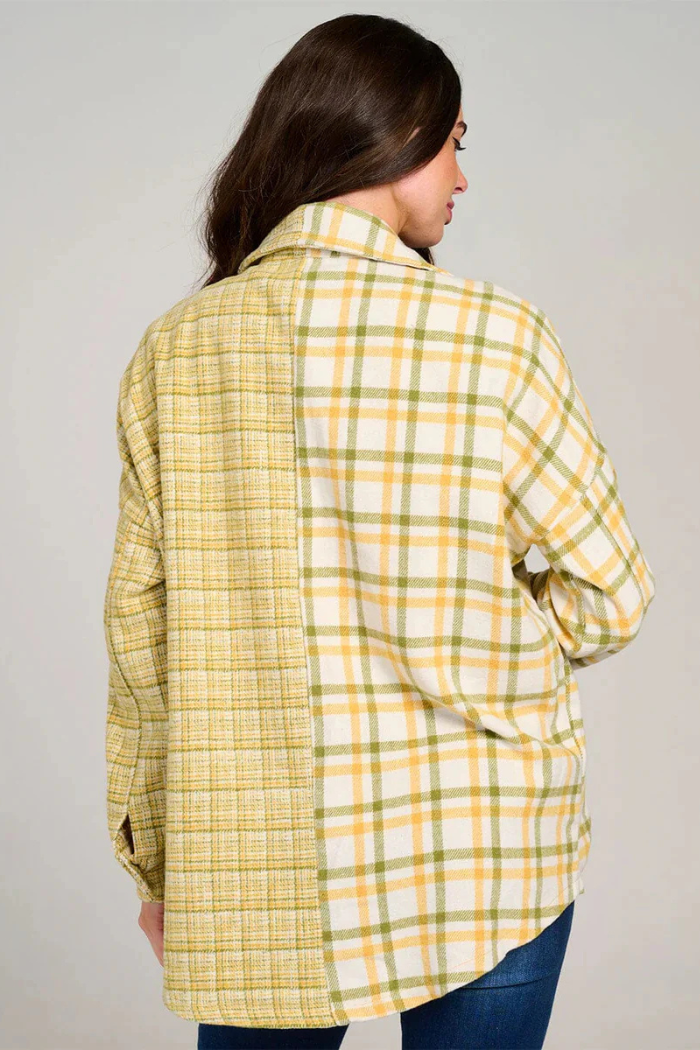 A woman is seen from behind, wearing a "shacket" or shirt-jacket with a distinctive mixed plaid pattern. The garment is split vertically down the back; the right half features a larger yellow, green, and white plaid, while the left half displays a smaller, tighter yellow and green plaid. She has dark hair and is wearing dark pants. 