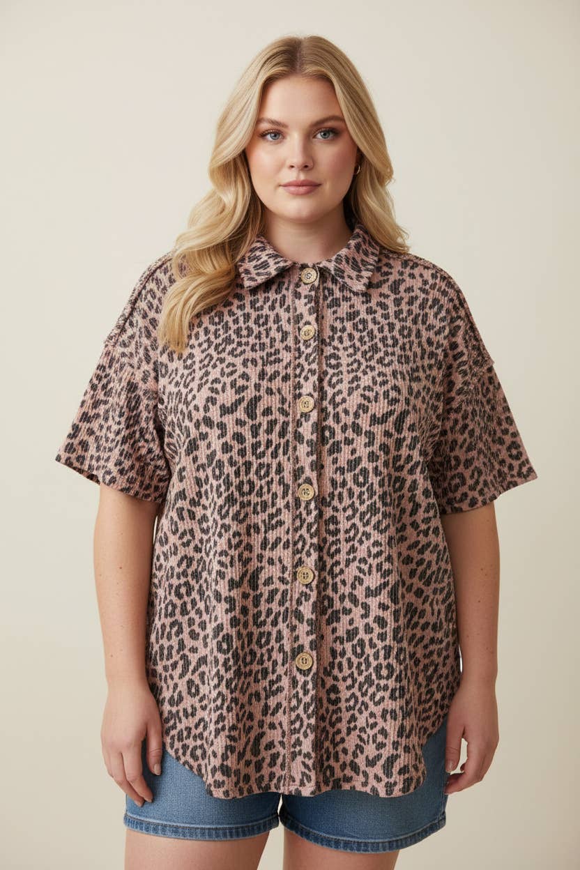 A woman with long blonde hair wears the Plus Leopard Button Blouse, a loose-fitting, short-sleeve, pink and black leopard print top paired with denim shorts, standing against a plain light background.