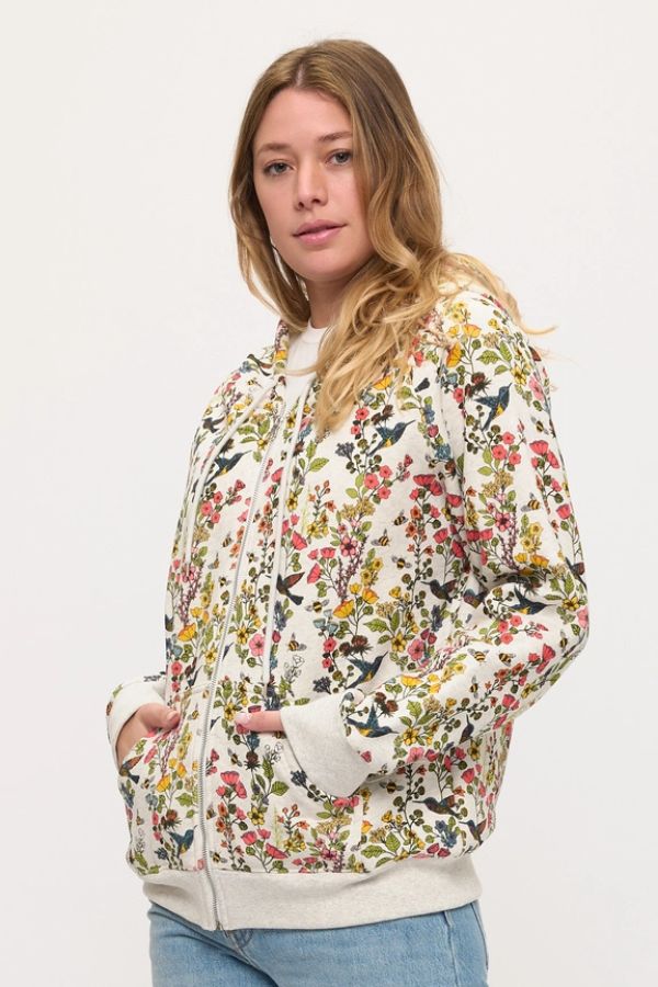 A woman with long hair stands casually with her hands in the pockets of a cream-colored Floral Hummingbird Zip Hoodie featuring a vibrant floral print. She pairs it with light blue jeans and looks at the camera, highlighting the hoodie’s relaxed fit.