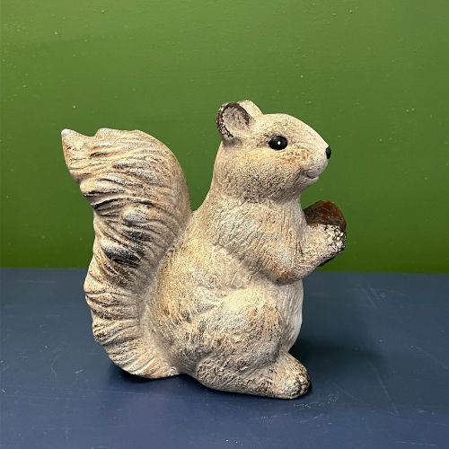 The Scamper Squirrel Figurine, a charming ceramic piece holding an acorn, sits on a blue surface with a green background, adding a touch of farmhouse décor that’s perfect for fall decorating.