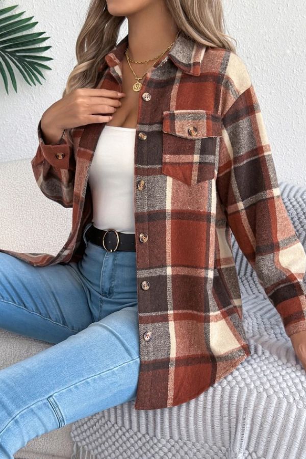 A woman sits on a couch wearing the Rust Plaid Longline Coat over a white tank top, paired with light blue jeans, a black belt, and layered gold necklaces. Her hand touches her collar.
