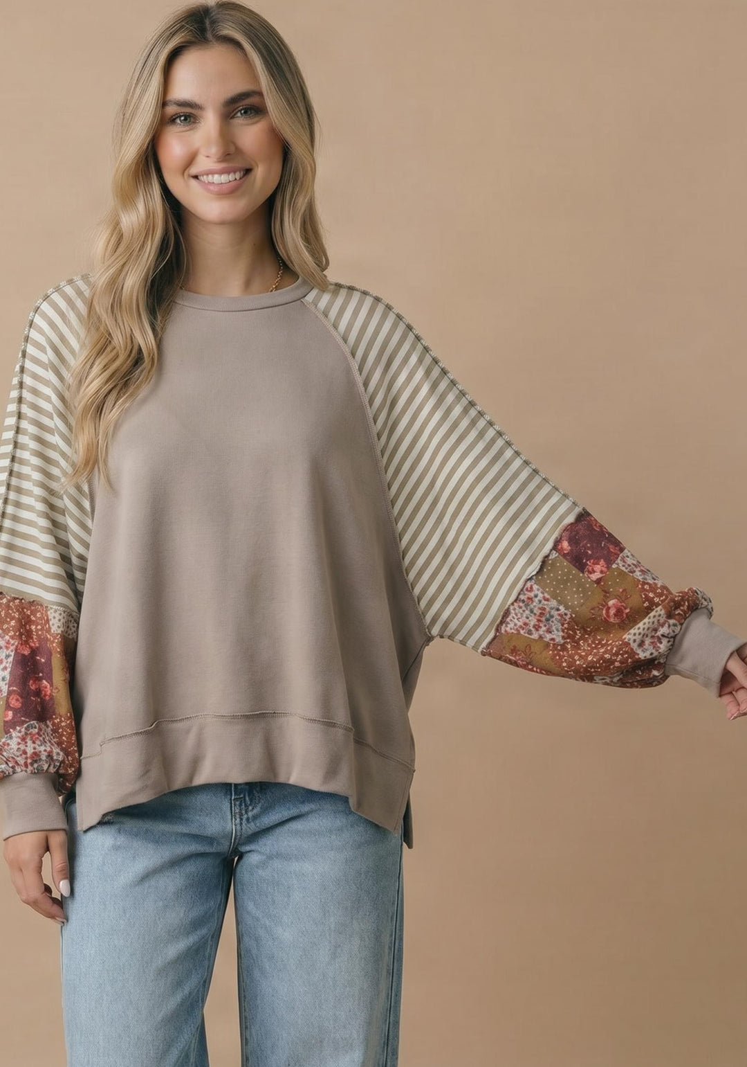 A woman with long, wavy blonde hair smiles at the camera in front of a beige background, wearing light blue jeans and the Patchwork Bubble Sleeve Top—a loose taupe sweatshirt featuring striped and floral patchwork bubble sleeves.