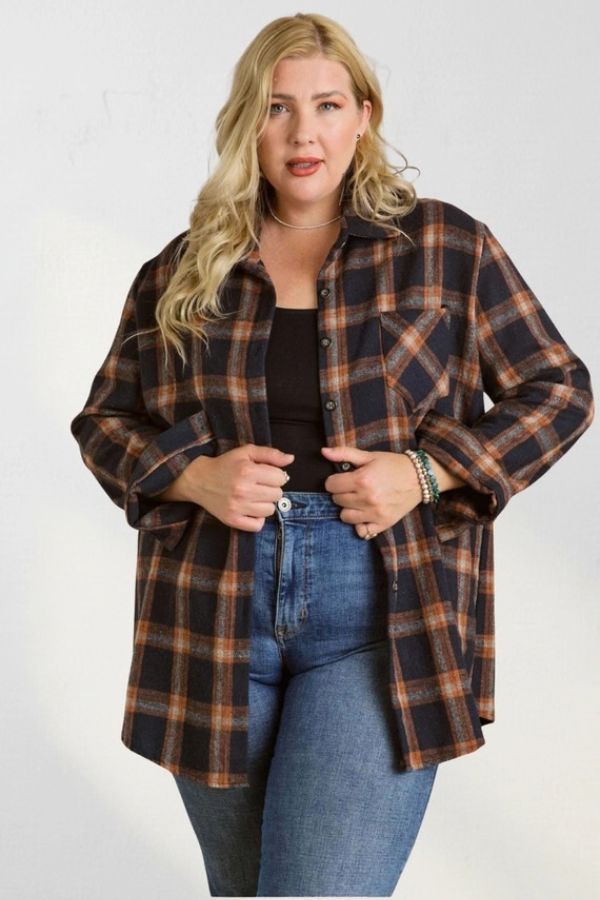 A woman wears a Curvy Plaid Flannel Shirt in brown and black plaid, unbuttoned over a black top and blue jeans.
