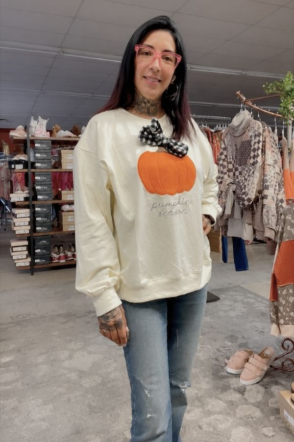 A woman wearing glasses, jeans, and the Pumpkin Season Bow Sweatshirt—a cream autumn top featuring an orange pumpkin and “pumpkin season” text—stands in a clothing store with racks and shelves of clothes and shoes in the background.