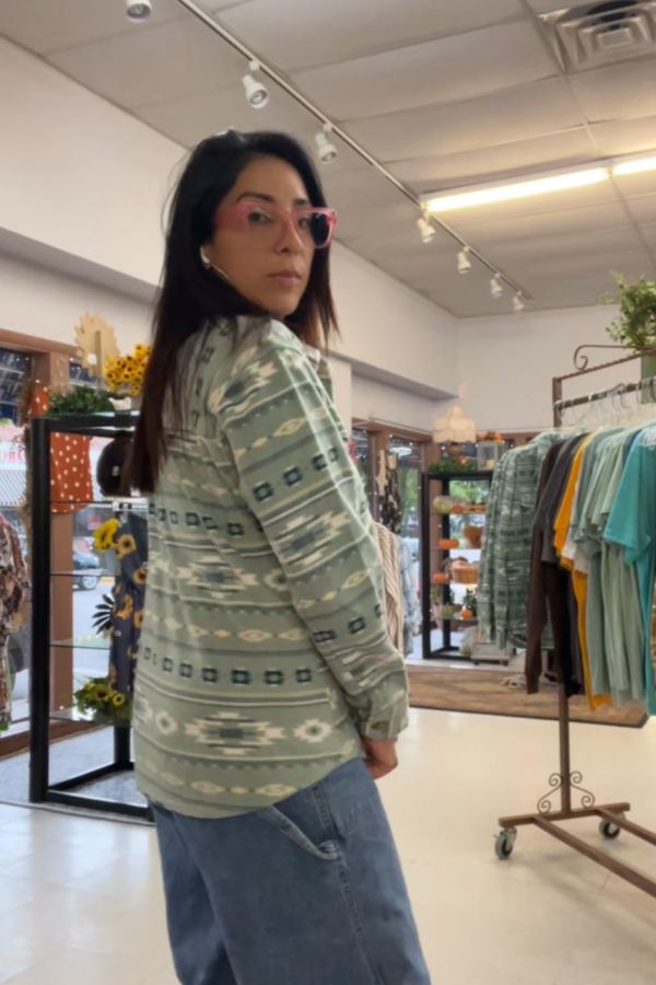 A person with long dark hair, wearing pink glasses, a patterned green shirt layered under the Southwestern Fleece Shacket, and jeans, stands in a clothing store with racks of clothes and plants visible in the background.