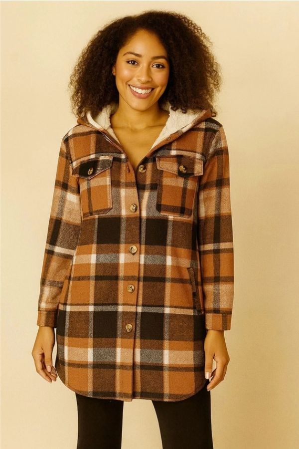 A woman with curly hair smiles at the camera, wearing the Fur Lined Plaid Hooded Shacket—a cozy, brown and black plaid button-up coat with front pockets—against a beige background.