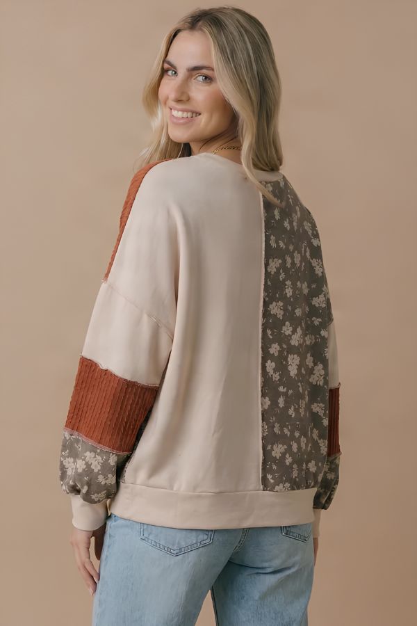 A woman with long blonde hair smiles over her shoulder while wearing the Color Block Knit Top, which features patchwork with floral and solid patterns in beige, brown, and rust tones. She pairs the top with light blue jeans against a beige background.