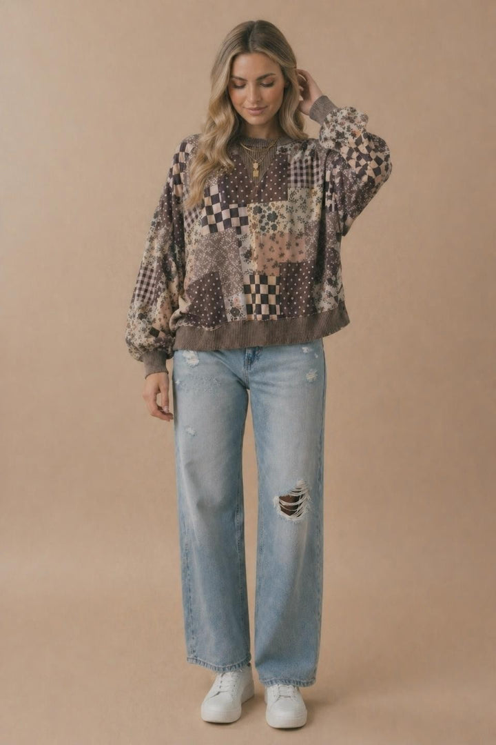 A person wearing the Plus Patchwork Pullover, paired with light ripped jeans and white sneakers, stands against a plain beige background, with one hand touching their hair and a relaxed expression.