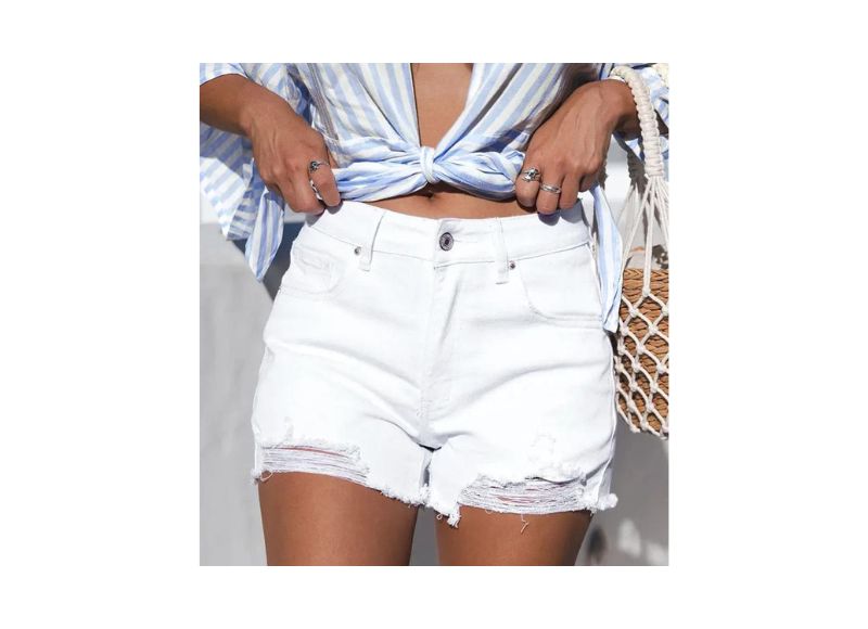 A woman wears a blue-and-white striped, tied-front blouse and White Distressed Denim Shorts with a raw hem, holding a woven straw bag. Only her midsection and hands are visible.