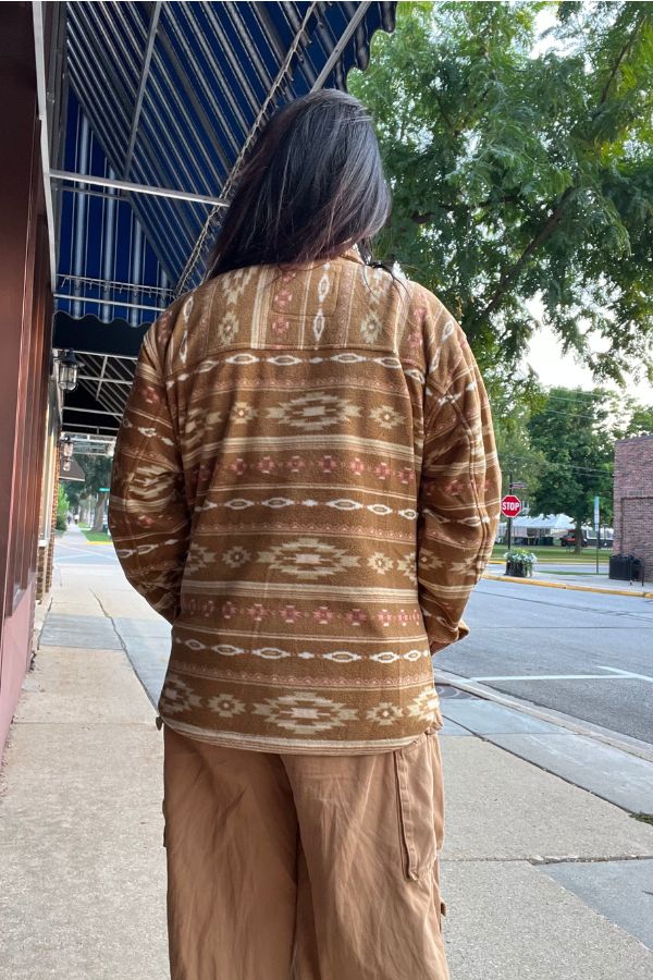 A person with long hair, wearing the Southwestern Fleece Shacket and tan pants, stands on a sidewalk facing away from the camera near a quiet street lined with trees and buildings.