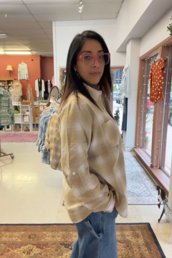 A woman wearing pink glasses, blue jeans, and the Plaid Button Shirt stands in a boutique store with clothing racks and home decor visible in the background.