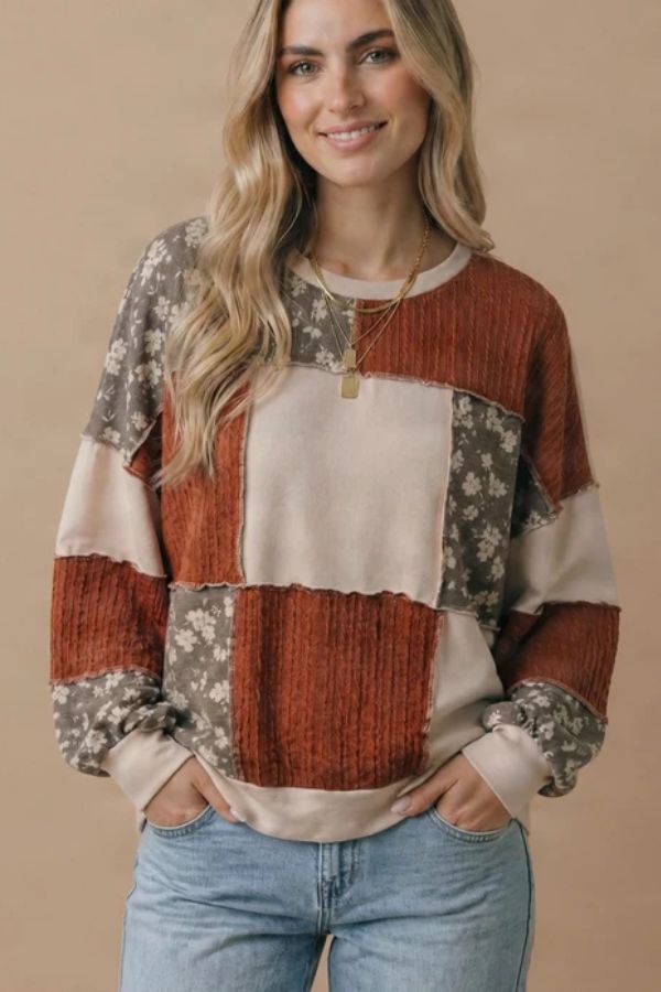 A woman with long, wavy blonde hair is wearing the Plus Color Block Top—a plus size top showcasing floral patchwork and color block details in rust, beige, and floral prints—paired with light blue jeans. She stands against a tan background, smiling with her hands in her pockets.
