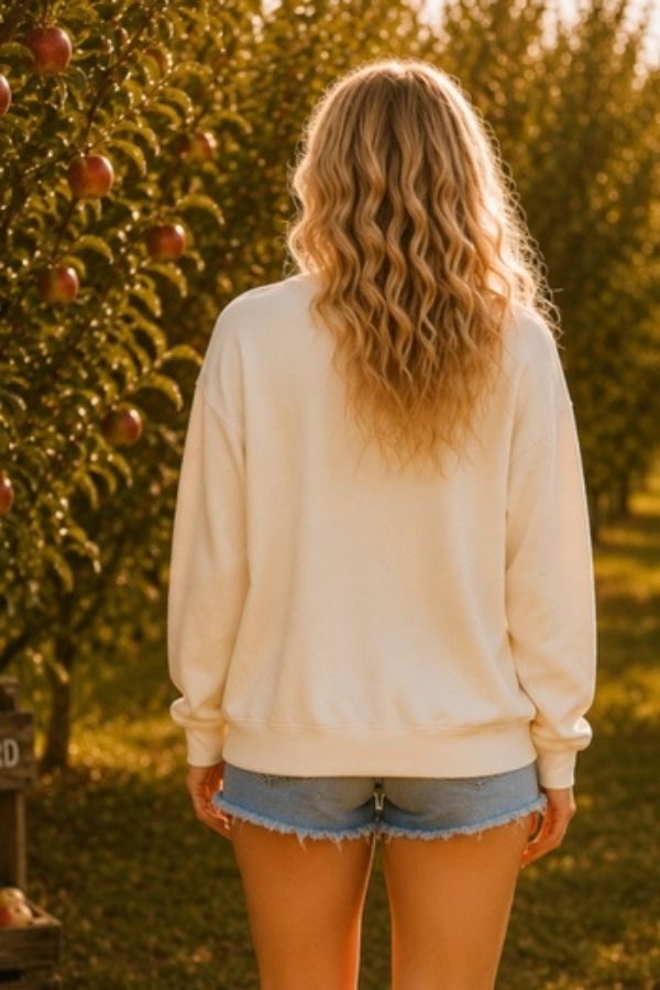 Wearing the Pumpkin Season Bow Sweatshirt and denim shorts, a person with wavy blonde hair stands in an apple orchard, facing away from the camera.