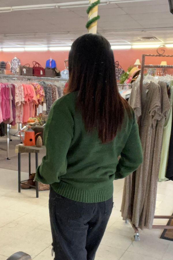A person with long, straight hair wearing the Sequin Reindeer Christmas Sweater and dark pants stands with their back to the camera in a clothing store filled with racks of clothes and accessories.