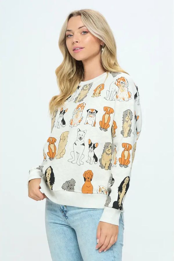 A woman with long blonde hair models the Dog Print Crewneck Sweatshirt, featuring a light-colored design adorned with various cartoon dog illustrations, paired with light blue jeans and set against a plain white background—perfect for dog lovers.