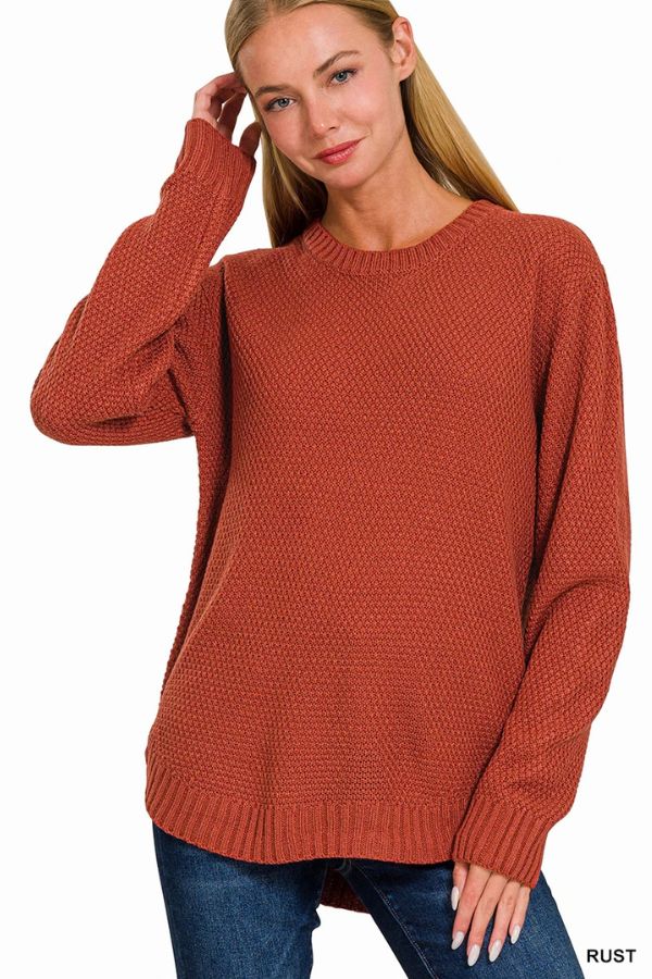 A woman wearing the Everyday Crew Neck Sweater in a rust shade and blue jeans stands against a white background, smiling gently with one hand touching her hair.