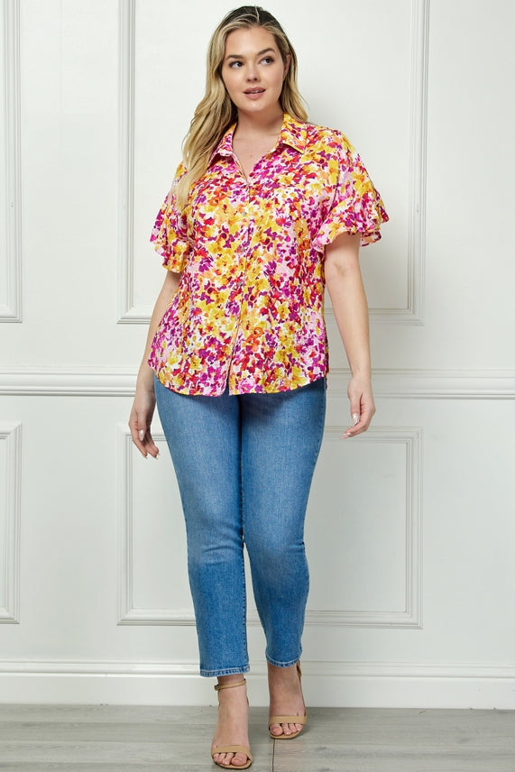A woman with long blonde hair stands confidently in front of a white paneled wall, wearing the Plus Floral Button Blouse—bright yellow and pink with flutter sleeves—paired with blue skinny jeans and beige heeled sandals.