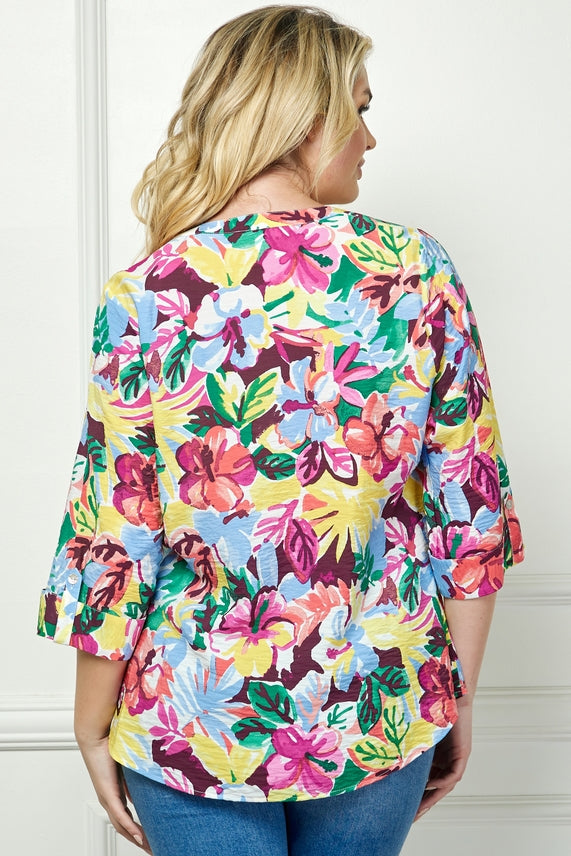 A woman with wavy blonde hair, seen from behind against a light-colored wall, wears the Plus Floral Placket Blouse—a relaxed fit blouse with a floral print and three-quarter sleeves.