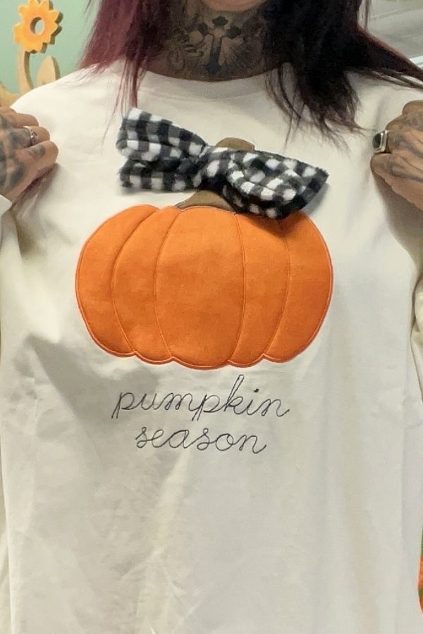 A person holds up the Pumpkin Season Bow Sweatshirt, a white autumn top featuring an orange pumpkin with a plaid bow and “pumpkin season” embroidered below it.