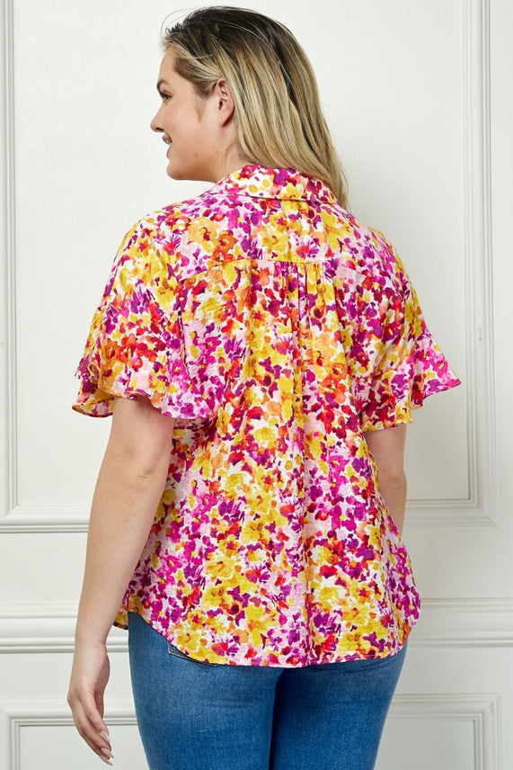 A woman faces away from the camera, wearing the Plus Floral Button Blouse—short-sleeve, loose-fitting with vibrant pink, yellow, and white flowers—paired with blue jeans, standing against a white paneled wall.