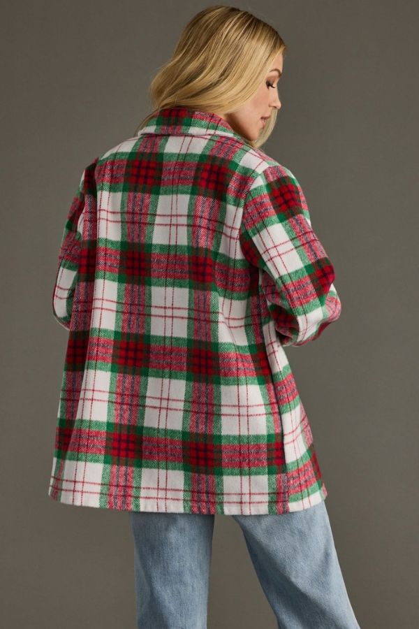 Wearing the Holiday Plaid Cozy Shacket—an oversized red, green, and white plaid jacket with a button-front closure—a person with long blonde hair stands against a plain gray background in light blue jeans, facing away from the camera for the perfect cozy fall look.