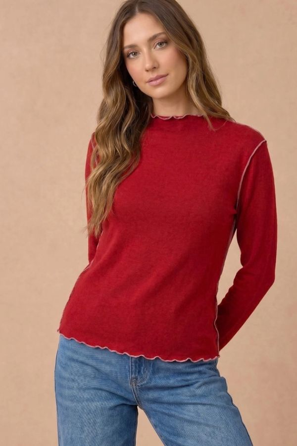 A woman wears a Frilled Mock Neck Hacci Top featuring a red mock neckline, long sleeves with lettuce-edge trim, visible light-colored seam detailing, and blue jeans.