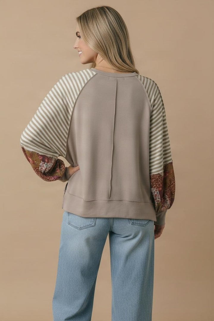 A woman faces away, wearing the Patchwork Bubble Sleeve Top—taupe French terry with striped and floral prints—paired with light blue jeans against a beige background.