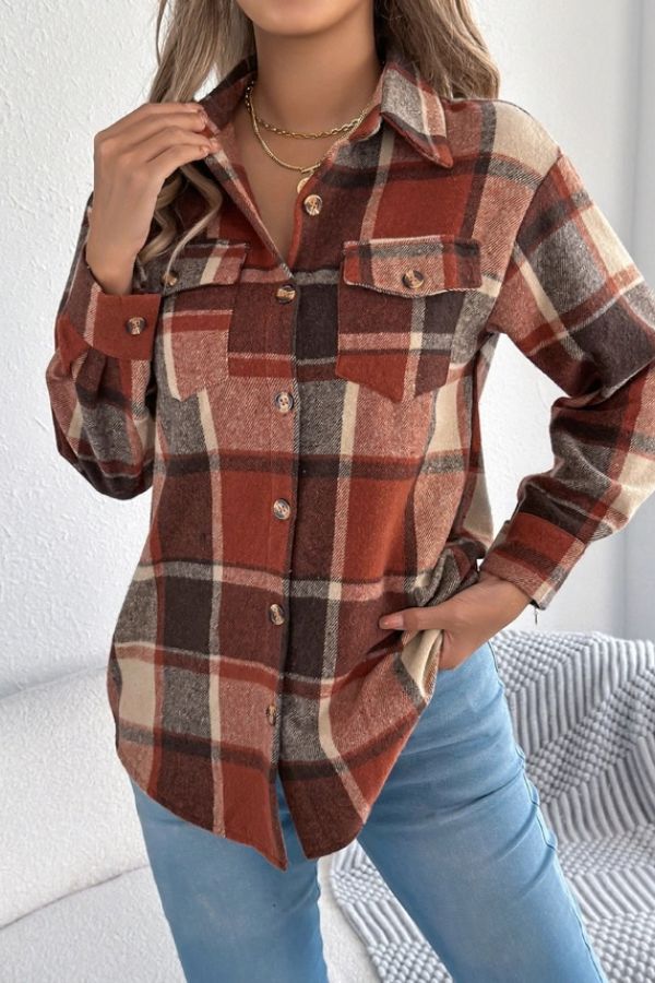 A woman wears the Rust Plaid Longline Coat featuring chest pockets and a brown, red, and beige plaid pattern, paired with light blue jeans. This lightweight layering piece is styled indoors, with one hand touching her collar and the other in her pocket.
