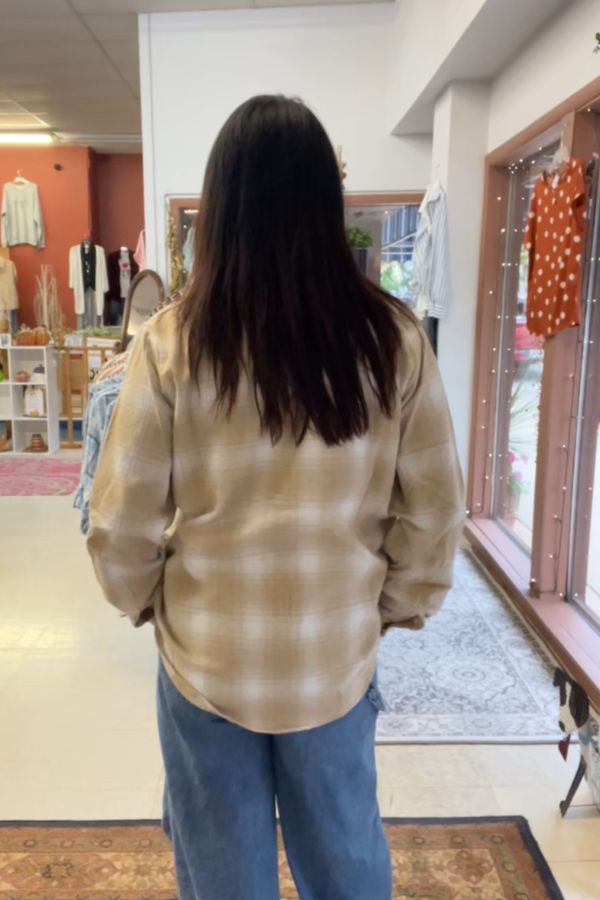 A person with long dark hair, wearing a beige Plaid Button Shirt and blue jeans, stands indoors facing away in a clothing store with racks, mirrors, and garments on display.