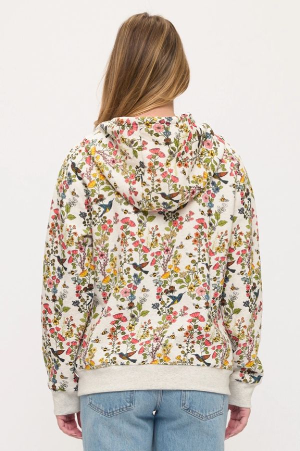 A person with light brown hair, dressed in the Floral Hummingbird Zip Hoodie—a relaxed fit cream zip hoodie featuring a colorful floral print—and blue jeans, is shown from the back against a plain background.