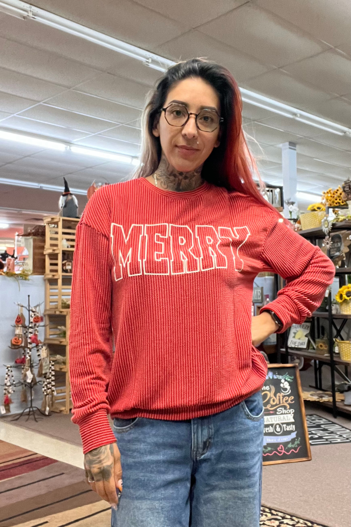 A woman wearing a red ribbed long-sleeve shirt with the word "MERRY" in white letters across the chest and blue jeans. She is posing with her left hand on her hip and her right hand resting on her side.