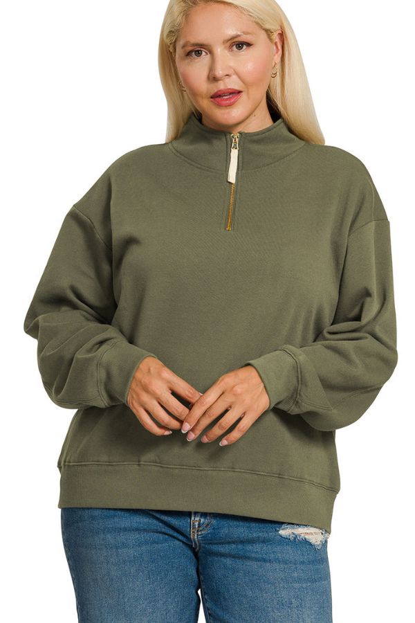 A person stands facing the camera with hands folded loosely in front, wearing the Plus Half Zip Sweatshirt in olive green, featuring a relaxed fit and high collar, paired with blue ripped jeans.