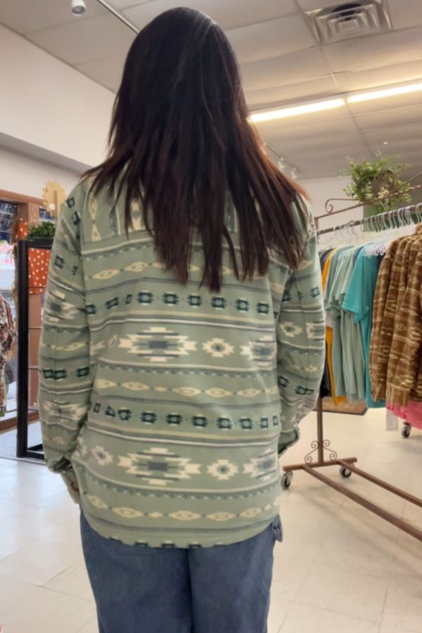 A person with long dark hair, dressed in a green patterned sweater and jeans, stands indoors facing away from the camera in a clothing store, browsing near a display of classic fleece shirts and Southwestern Fleece Shackets.