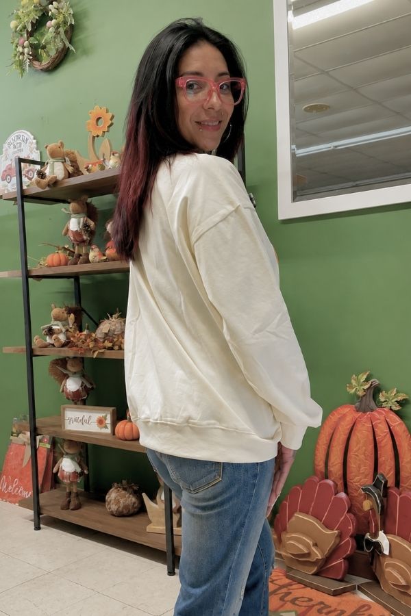 A woman with dark hair and pink glasses is smiling at the camera while wearing the cozy Pumpkin Season Bow Sweatshirt and jeans, standing indoors near autumn-themed decorations against a green wall.