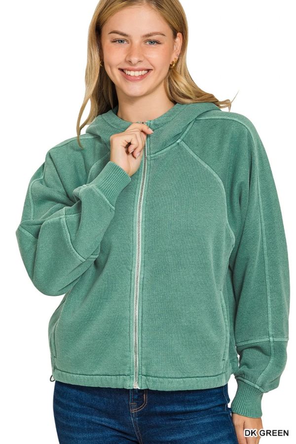 A smiling woman with blonde hair wears the Pigment Dye Zip Up Hoodie in light green and blue jeans. She holds the hoodie near her neck with one hand against a plain white background.