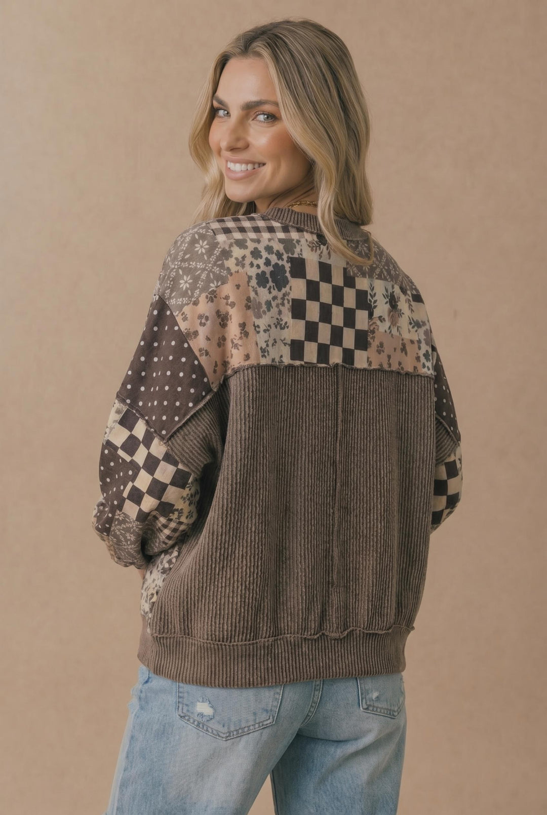 A woman smiles over her shoulder while wearing the Patchwork Oversized Pullover in brown and beige with assorted patterns, paired with light blue distressed jeans, standing against a plain beige background.