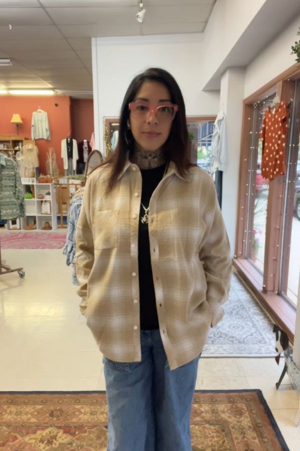 A woman with dark hair and glasses stands indoors, wearing the Plaid Button Shirt over a black top and blue jeans. She has tattoos on her neck and hands, and is standing in a boutique with racks of clothes and decor behind her.