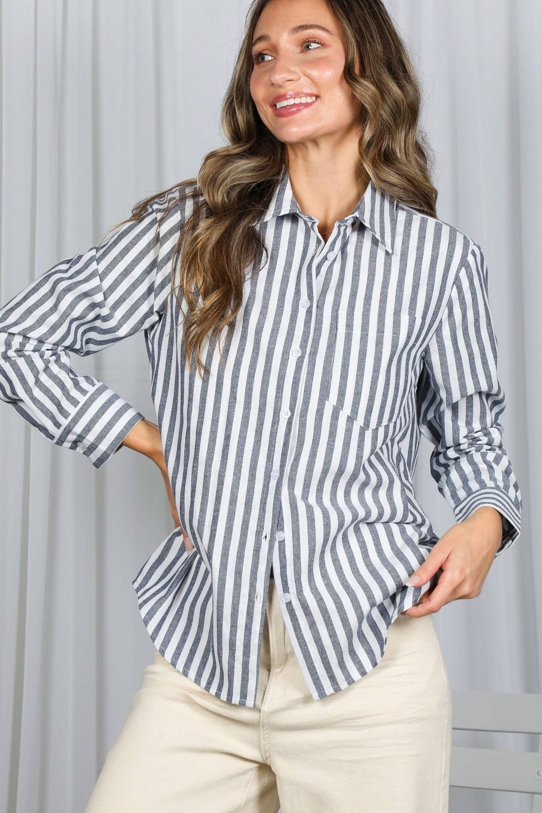 A woman smiles and poses with one hand on her hip, wearing the Striped Button Shirt featuring black and white vertical stripes along with light beige pants, as she stands in front of white curtains.