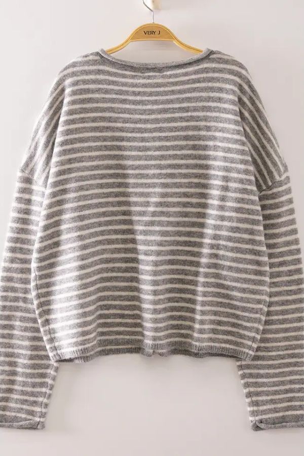 The Striped Button Front Sweater, featuring gray and white stripes, hangs on a wooden hanger against a light-colored wall, highlighting its cozy knit texture and relaxed fit.