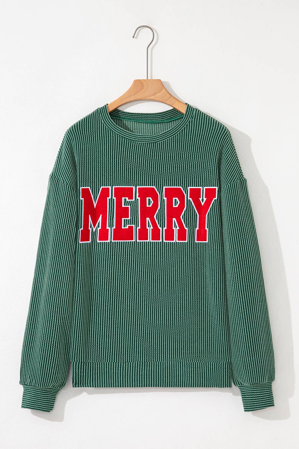 The Merry Graphic Top is a green ribbed sweater with long sleeves, shown hanging on a wooden hanger. Featuring the word "MERRY" printed in bold red and white capital letters across the chest, it's the ideal festive graphic top for winter wear and holiday gatherings.