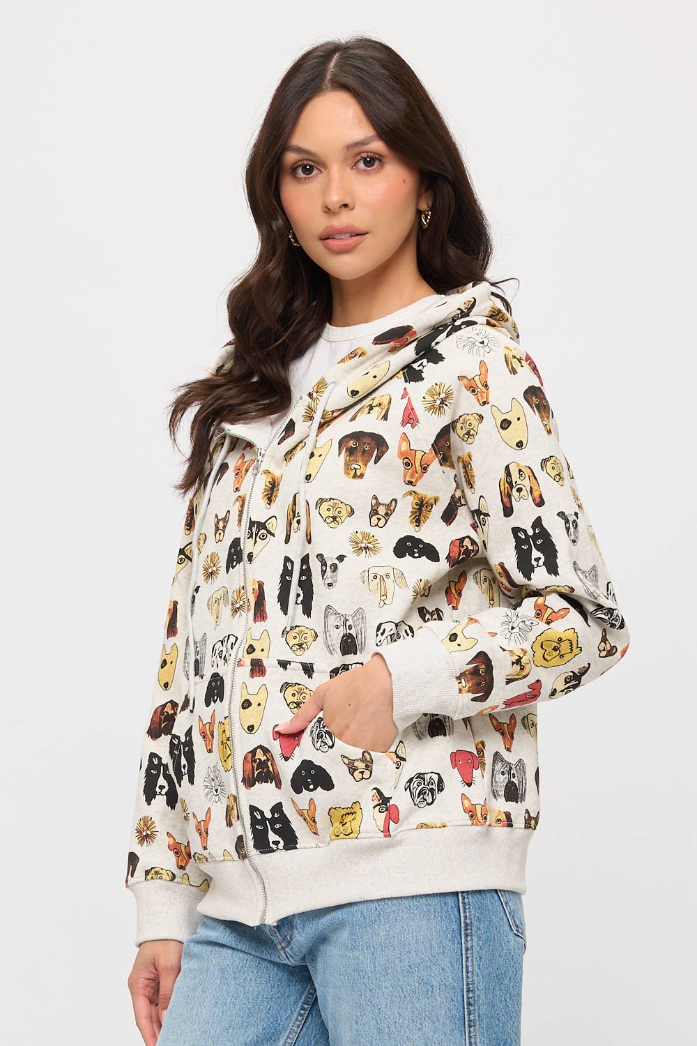 A woman with long dark hair models the COLORFUL DOG FACES PRINT ZIP UP HOODIE, featuring playful dog illustrations, with one hand in her pocket against a white background—a fun choice for any dog lover's wardrobe.