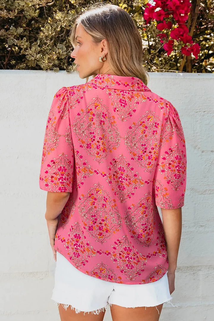 A woman with light hair stands outdoors facing away, wearing the Floral Puff Sleeve Blouse in pink with floral patterns and white shorts; lush greenery and bright pink flowers fill the background.