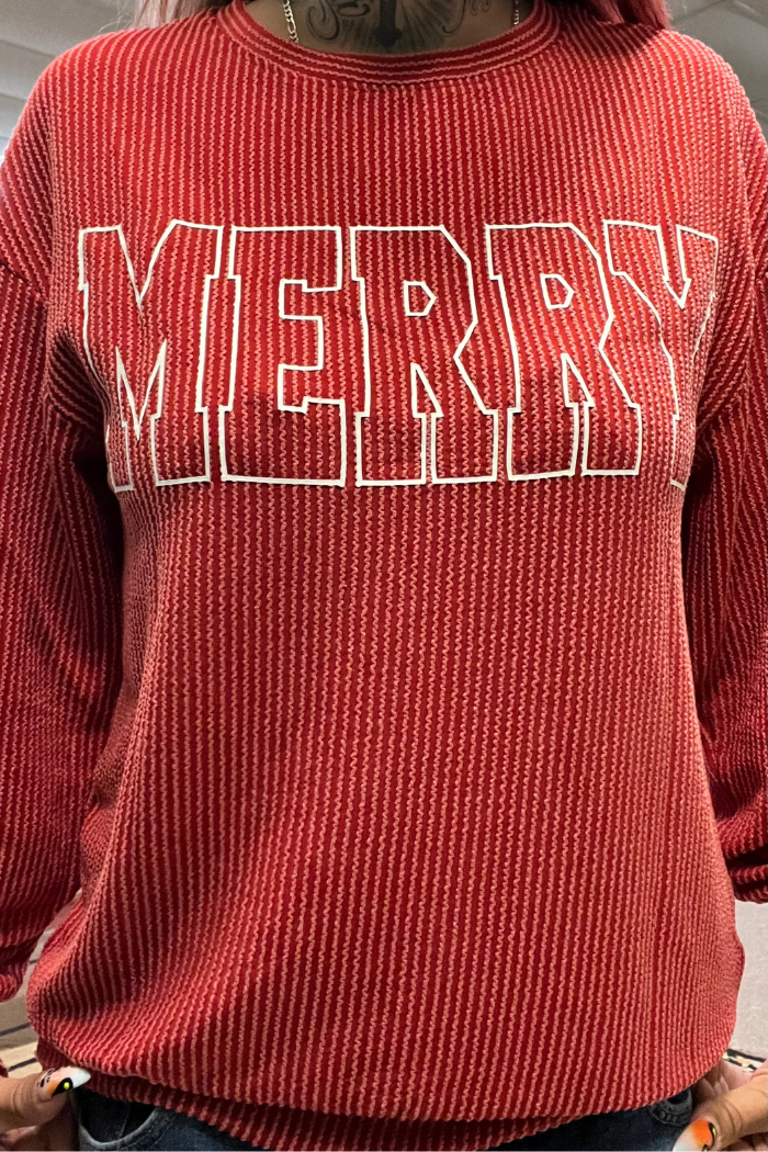 A close-up shot of a person wearing a red ribbed long sleeve shirt. The word "MERRY" is written in large, white, block letters across the chest.