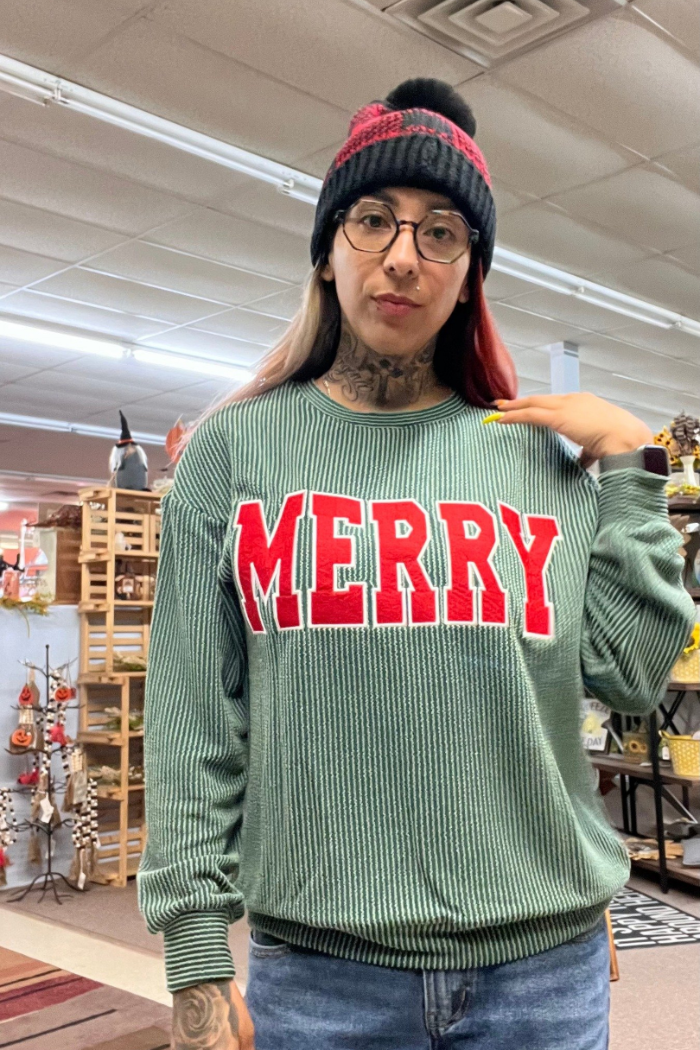 A woman with tattoos on her neck and arms is wearing a green, ribbed long-sleeve top with the word "MERRY" in large red letters across the chest. She is also wearing a black and red beanie and glasses.